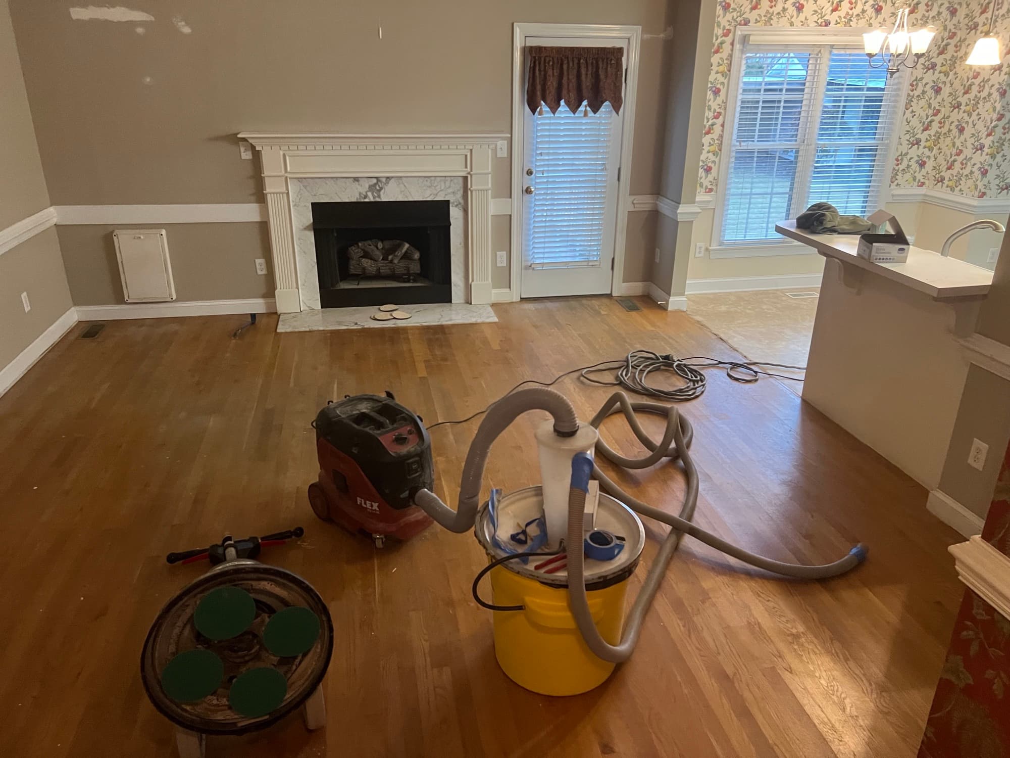 Sanding equipment staged in living room during red oak refinishing in Wilson NC, Festool vacuum and sanding discs visible, marble fireplace in background