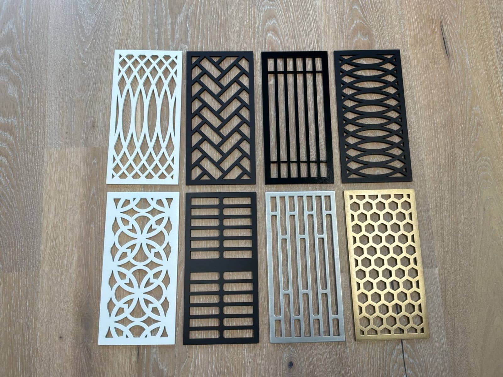 Eight quarter-inch steel flush-mount floor vent patterns in white, black, silver, and gold laid on a white oak floor